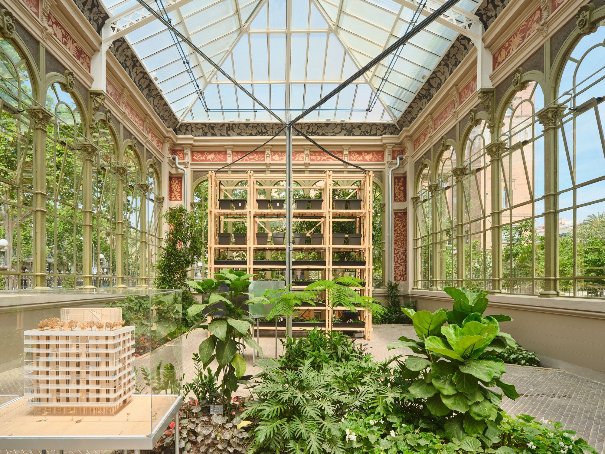 The model in front of the vertical planting system depicts Terrazas para la Vida, an 8-story timber social housing building in Barcelona designed by IAAC affiliates, which hosts a timber greenhouse on its rooftop for use by the residents.